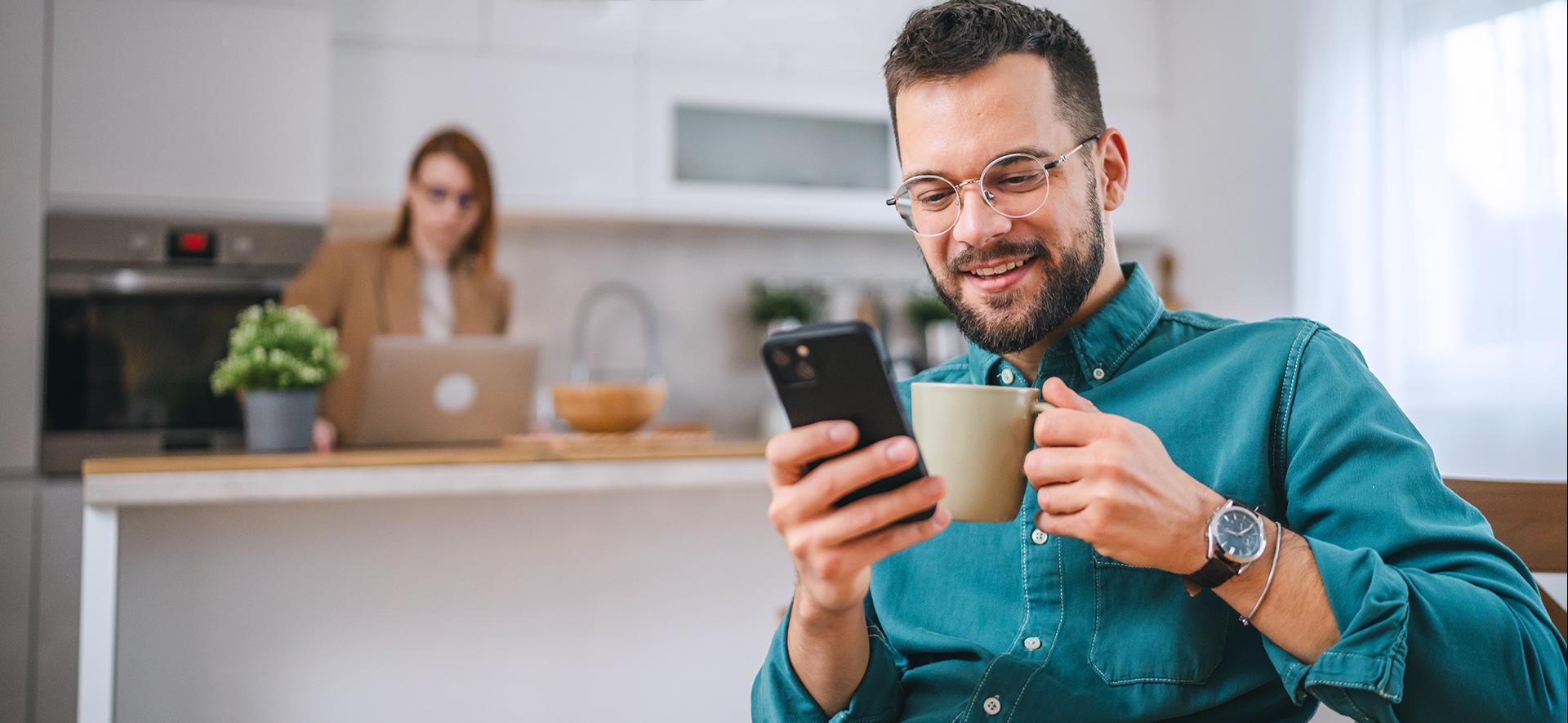 Uśmiechnięty mężczyzna z kubkiem w dłoni i telefonem, coś klikający.