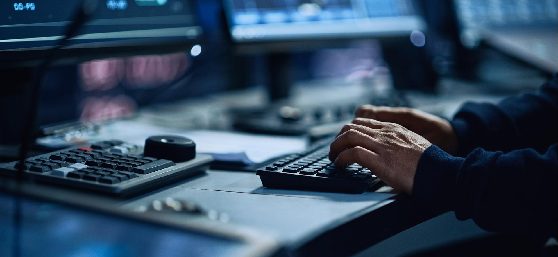 ręce mężczyzny piszącego na klawiaturze, w tle monitory